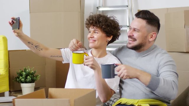 Panning waist up shot of pleased smiling Caucasian husband and wife making memorable selfies on smartphone while relaxing and drinking tea