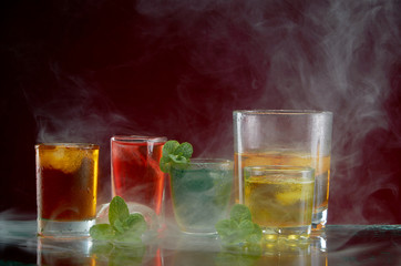 Different alcoholic cocktails with ice and mint with light smoke on a red background