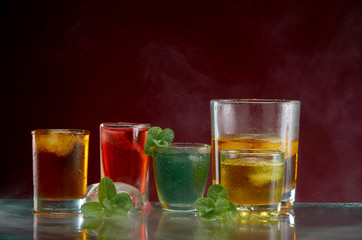 Different alcoholic cocktails with ice and mint with light smoke on a red background