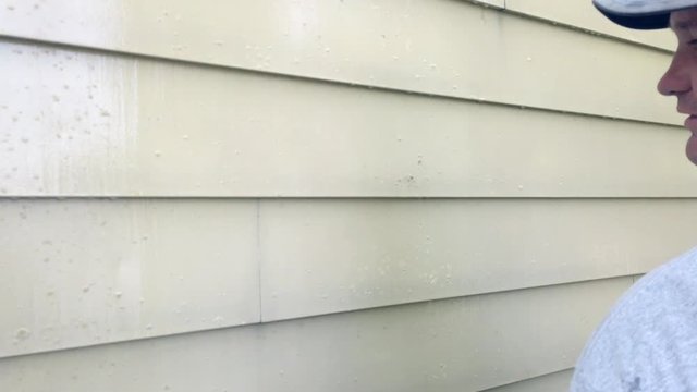 Man looking at the yellow siding on a house. He then tries to scrape of some of the dirt with his fingernail