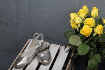 A large bouquet of yellow roses in a glass vase. Nearby is a wooden box. On the box is a pair of women's shoes of silver color.