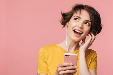 Happy young beautiful woman posing isolated over pink wall background listening music with earphones using mobile phone.