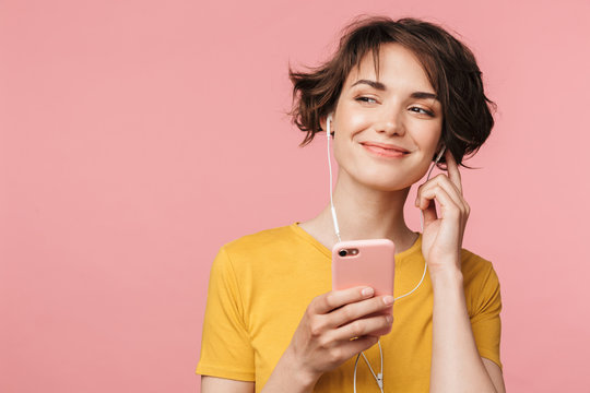 Happy young beautiful woman posing isolated over pink wall background listening music with earphones using mobile phone.