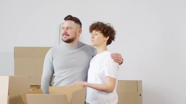 Panning medium shot of excited Caucasian couple standing in loving embrace in their new home, admiring it and discussing future interior refurbishment and furniture arrangement