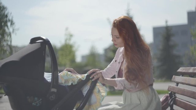 Young happy redhead mother sitting on bench near the house with baby carriage and smiling and lulling him on a nice spring day