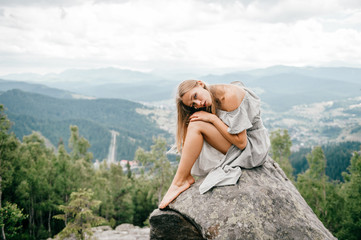 Naklejka premium Lonely young girl sits at stone on top of mountain with far view at hills and cloudy sky and hugs her legs with head lying on knees. Beautiful blonde woman outdoor summer portrait. Female at nature.