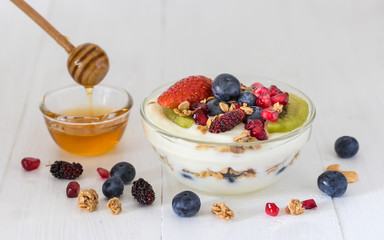 Plain yogurt with strawberry, blueberries, kiwi, granola, pomegranate in a glass bowl and honey on white wooden texture, healthy food and plant-based food concept