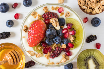 Top view of plain yogurt with strawberry, blueberries, kiwi, granola, pomegranate in a glass bowl and honey on white wooden texture,flay lay of healthy food and plant-based food concept
