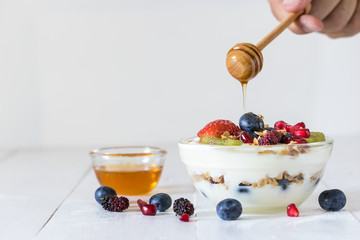 Plain yogurt with strawberry, blueberries, kiwi, granola, pomegranate in a glass bowl and honey on white wooden texture, healthy food and plant-based food concept