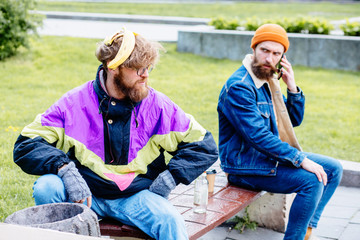Homeless man with banana skin near full garbage basket in colorful clothes rummaging in trash container in park green background. Shocked hansome beard male sitting on the bench near him