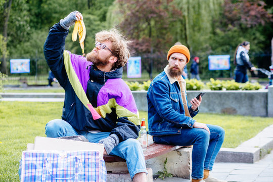 Homeless man with banana skin near full garbage basket in colorful clothes rummaging in trash container in park green background. Shocked hansome beard male sitting on the bench near him