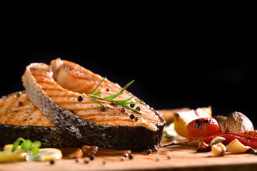 Grilled salmon fish and various vegetables on wooden table on black background