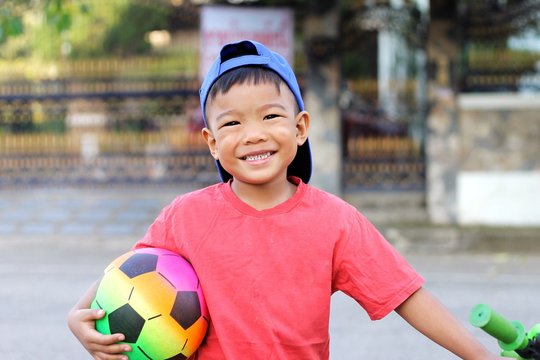 At the playground. Happy asian child boy smiling and holding a ball toy in his hand. He wearing a red shirt and blue cap. Kid and sport concept.