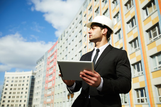 Low angle portrait of handsome Middle-Eastern businessman wearing hardhat standing against apartment building on construction site, copy space