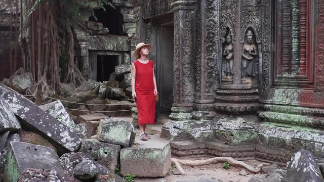 Caucasian woman in red dress is walking among ruins of beautifully decorated Ta Prohm temple and taking photos. Angkor Wat complex. Cambodia