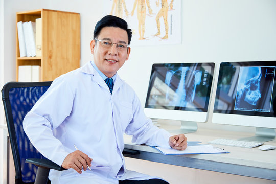 Portrait of Asian male doctor sitting in white coat at the table in front of computer monitors with x-ray images and looking at camera