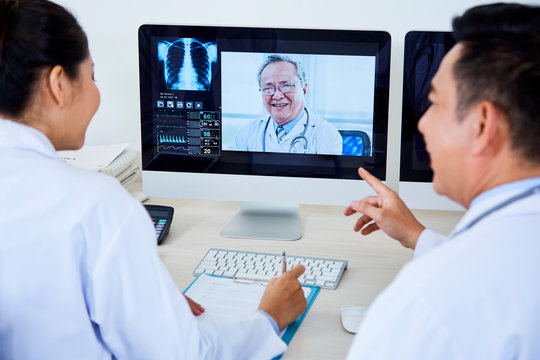 Rear view of two Asian doctors sitting in front of computer monitor and discussing with their colleague working moments during online conference