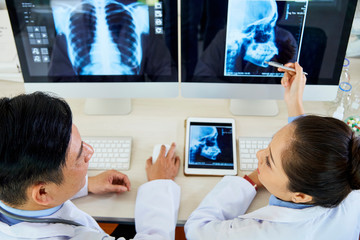 Two Asian radiologists sitting at the table in front of computer monitors with x-ray images and...