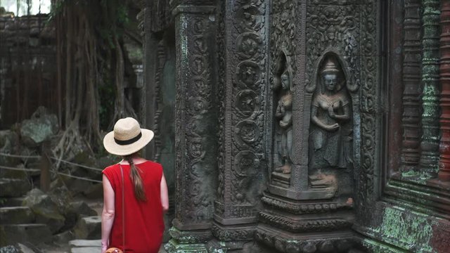 Caucasian woman in red dress and straw hat is walking among ruins of beautifully decorated Ta Prohm temple in Angkor Wat complex. Cambodia