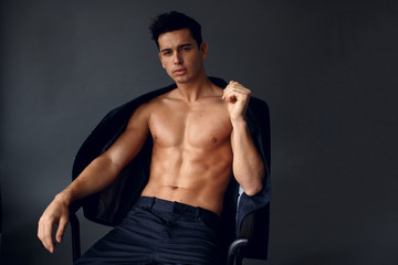 Handsome, shirtless young man with jacket on his shoulders, seated on chair, on grey background.