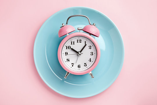 Pink larm clock on empty blue plate. Concept of intermittent fasting, lunchtime, diet and weight loss