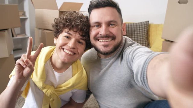 Chest-up shot of delighted middle-aged Caucasian couple sitting on floor and taking selfies on smartphone against cardboard boxes in their new home after house move, smiling, waving and doing v-sign