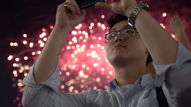 Asian man takes the phone fireworks. Japanese in a shirt takes on the phone salute. A man shoots a video on a phone vertically close-up. Holiday recording on a smartphone at night. Boy takes pictures