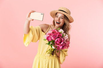 Beautiful amazing young blonde woman posing isolated over pink wall background holding flowers using mobile phone take a selfie.