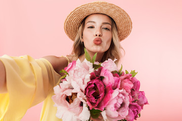 Amazing young blonde woman posing isolated over pink wall background holding flowers take a selfie by camera.