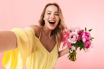 Amazing young blonde woman posing isolated over pink wall background holding flowers take a selfie...