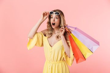 Beautiful shocked young blonde woman posing isolated over pink wall background holding shopping...