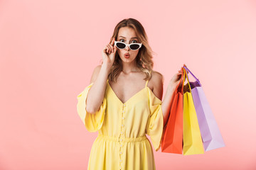 Beautiful shocked young blonde woman posing isolated over pink wall background holding shopping bags.