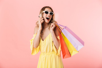 Beautiful excited happy young blonde woman posing isolated over pink wall background holding shopping bags talking by mobile phone.