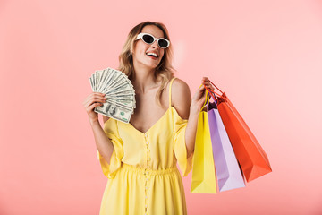 Excited happy young blonde woman posing isolated over pink wall background holding shopping bags and money.