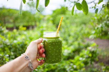 woman's hand holding jar with green cold-pressed juice, vegetable garden background. Healthy...