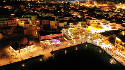 Aerial drone night shot of iconic and picturesque illuminated Venetian old port of Chania with famous landmark lighthouse and shops - restaurants serving Cretan traditional food, Crete island, Greece