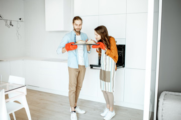 Portrait of a young beautiful couple holding tray with fresh baked breads in the modern bright kitchen at home