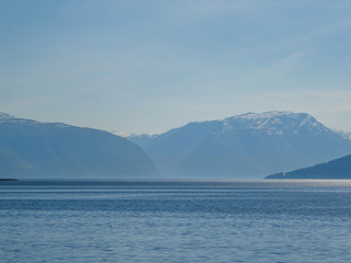 A view on the Songefjorden (King of the Fjords) from the water level. It is the deepest fjord in Norway. Tall, lush green mountains surrounding the fjord. Calm surface of the water. Clear blue sky.