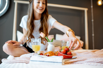 Young and cheerful woman having breakfast while sitting on the bed at cozy bedroom