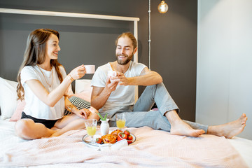 Young and cheerful couple eating breakfast, sitting together on the bed at home