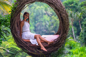 Beautiful tan woman in white dress posing on a straw nest. Rain forest on the background