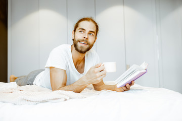 Handsome man in pajamas reading book while relaxing with coffee on the bed at home