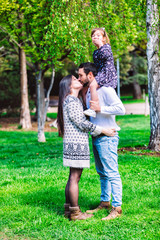 Fototapeta premium Family mom, dad and daughter sits at the daddy on the shoulders, and parents kiss on the park at daylight