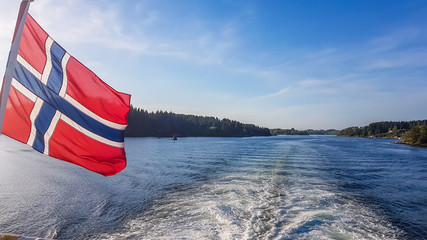 Norwegian flag hanging on  the railing of the ship and waving above the water.The motor of the ship makes the water wavy and foamy. Tall, lush green mountains surrounding the fjord. Clear blue sky.