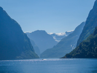 A view on the Songefjorden (King of the Fjords) from the water level. It is the deepest fjord in Norway. Tall, lush green mountains surrounding the fjord. Calm surface of the water. Clear blue sky.