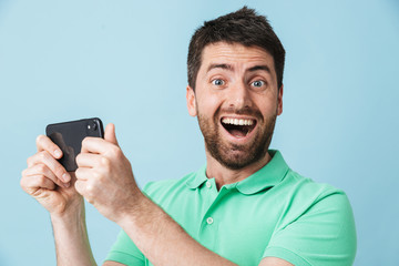 Excited emotional young handsome bearded man posing isolated over blue wall background play games by mobile phone.