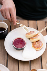 Breakfast with friends in a summer cafe in the open air. On the table are dessert fried cottage cheese pancakes with red jam, orange juice and black coffee. Hands in the frame with golden cutlery.