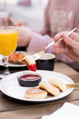 Breakfast with friends in a summer cafe in the open air. On the table are dessert fried cottage cheese pancakes with red jam, orange juice and black coffee. Hands in the frame with golden cutlery.