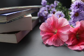 Three petunia flowers are in a vase that is on a stack of books. Nearby is an open diary and a white pen. Gray stone background.