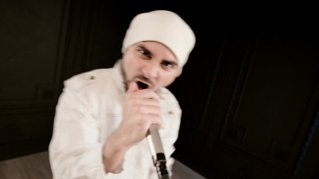 Frontman vocalist rock pop with a stylish beard in white clothes and a hat with a microphone in his hands expressively aggressively singing in the studio against the background of black walls
